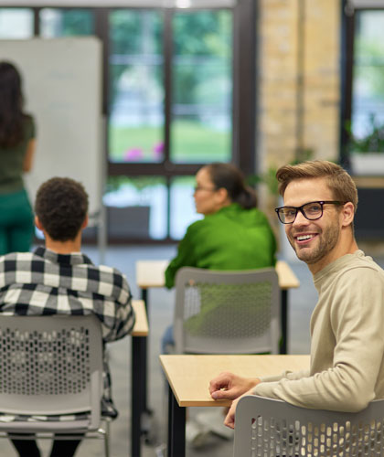 Hombre con gafas sonriendo sentado en un aula, con otras personas en mesas al fondo.
